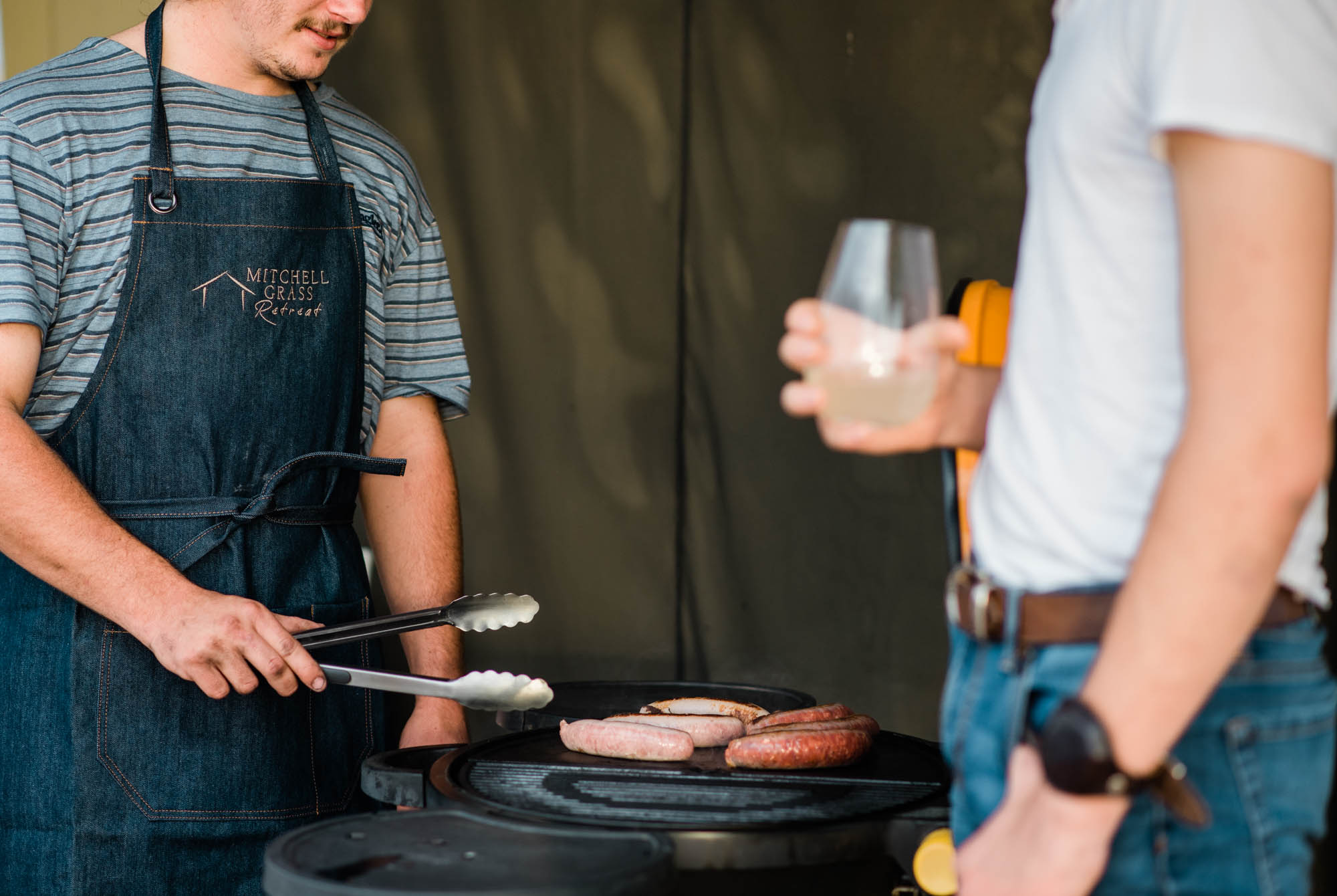 Man with tongs cooking sausages on a BBQ, wearing a denim apron with embroidered Mitchell Grass Retreat logo.